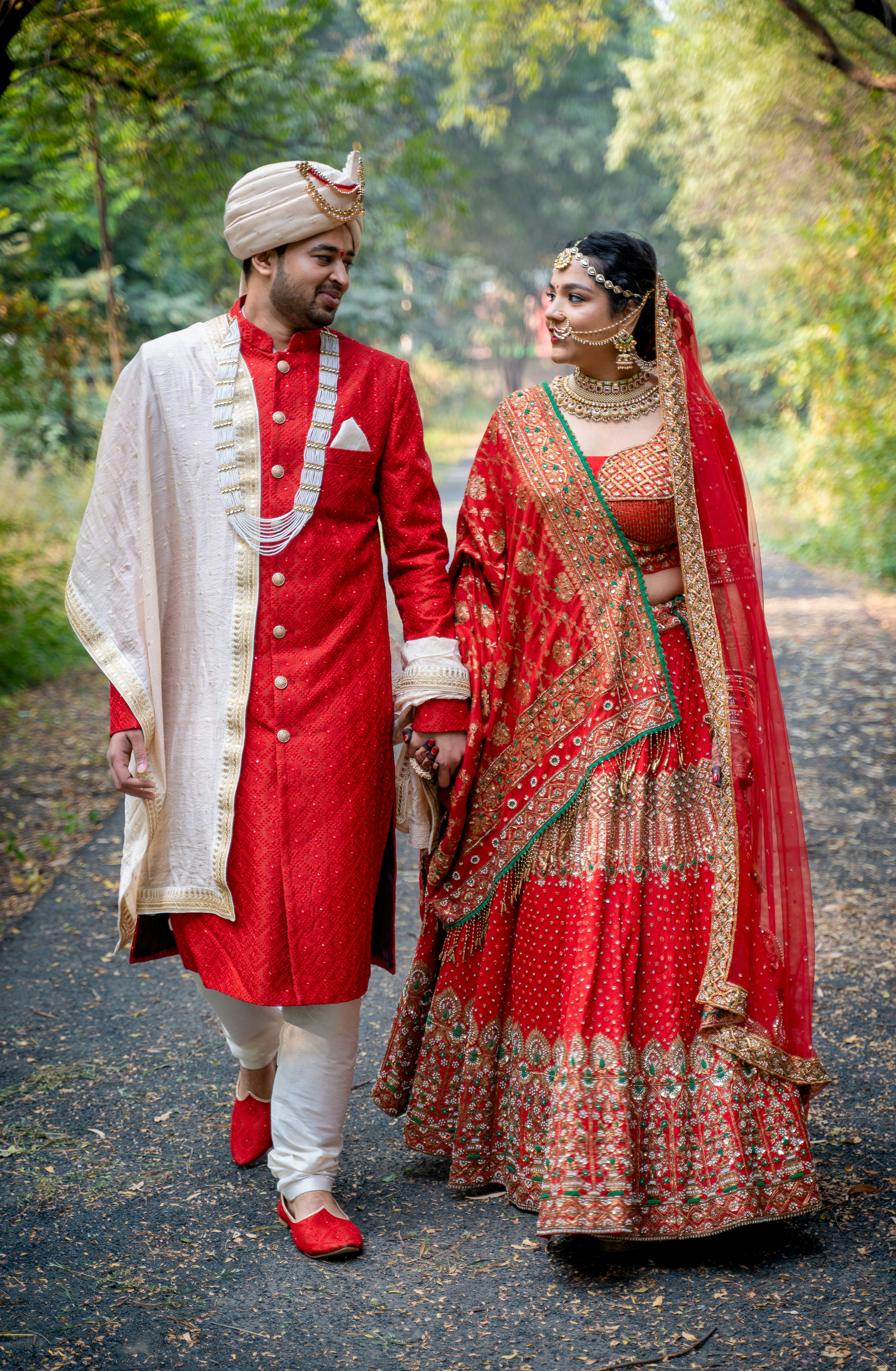 Couple in red bridal attire walking hand in hand along a tree-lined avenue