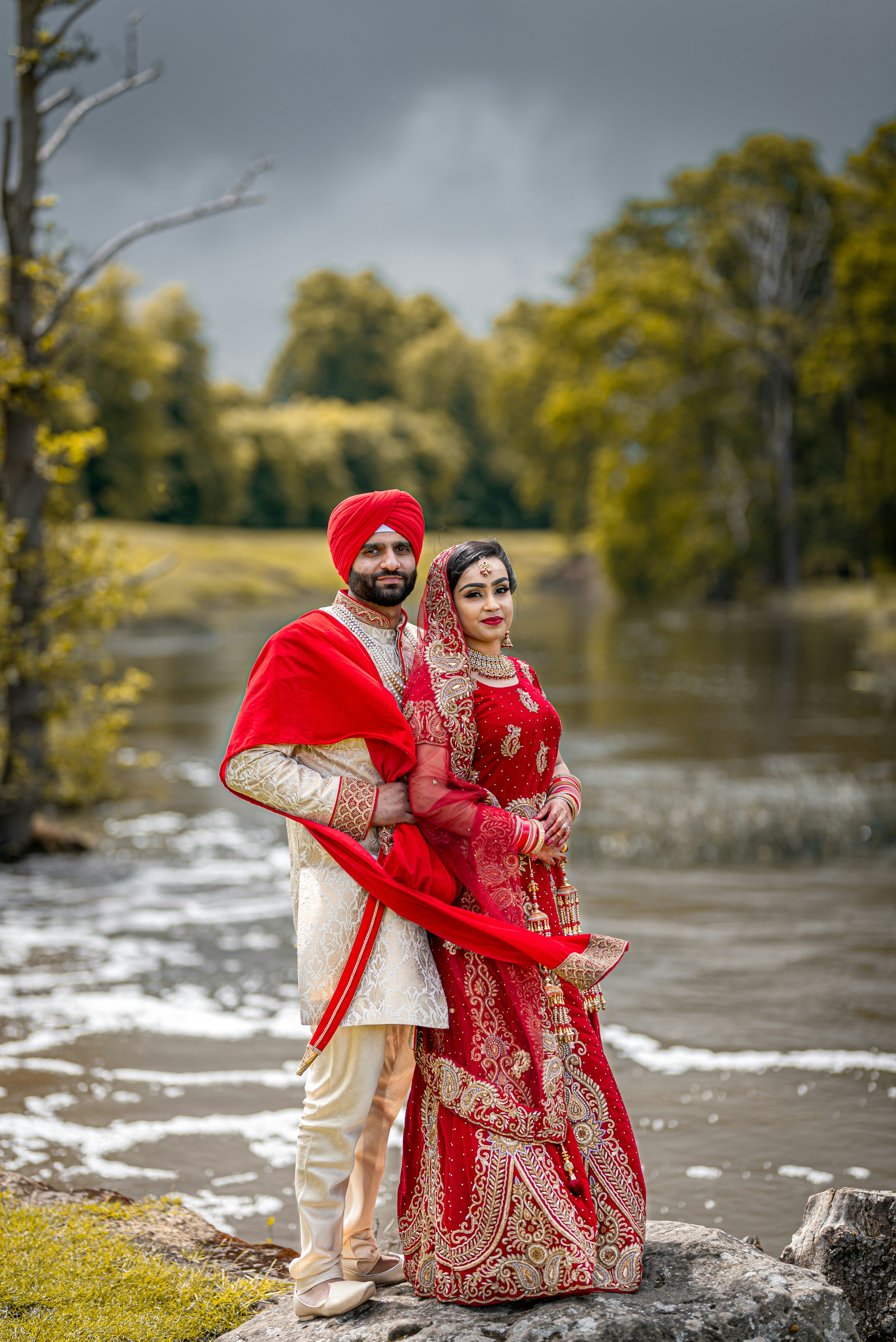 Sikh couple in red bridal attire standing by a river under a dramatic sky